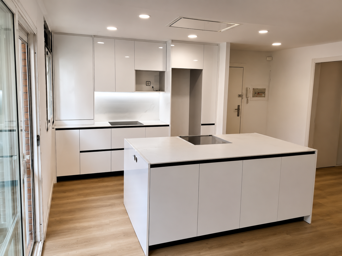 Cocina blanca abierta con gran isla central, encimera clara, muebles sin tiradores y columnas de almacenaje