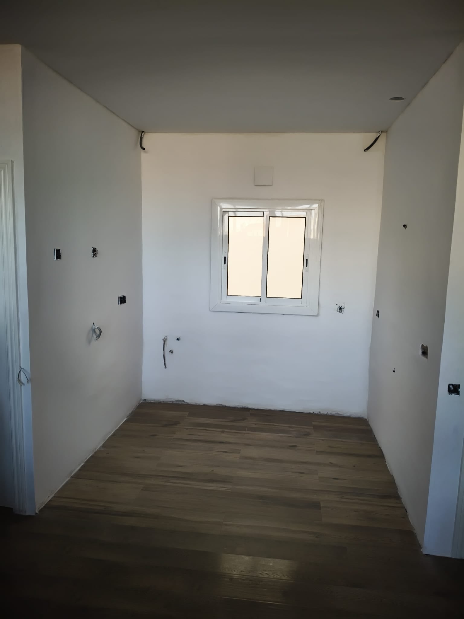 Espacio estrecho y vacío antes de instalar una cocina, con ventana, suelo efecto madera y conexiones preparadas